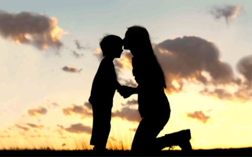 Silhouette of a kneeling mother kissing her son on the forehead in front of a sunset.