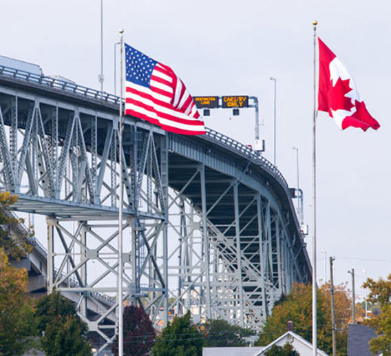 Us and Canada flag waving with border checkpoint in background 