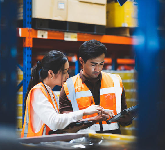Man and woman in orange vests looking at a tablet inside a warehouse