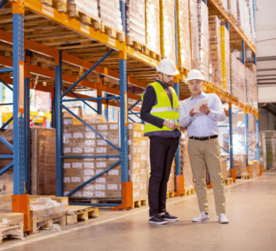 Two men in hardhats look over paperwork in a warehouse.