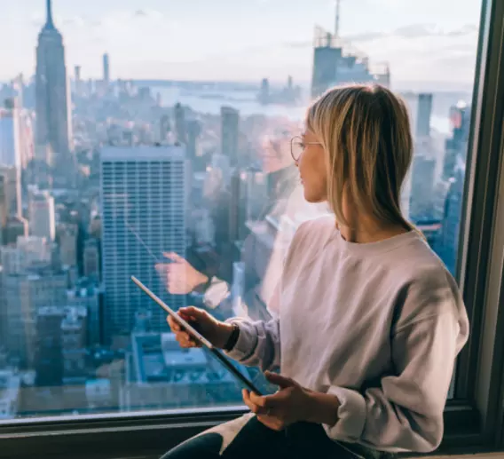 Woman holds a tablet and looks out a window overlooking the New York City skyline.