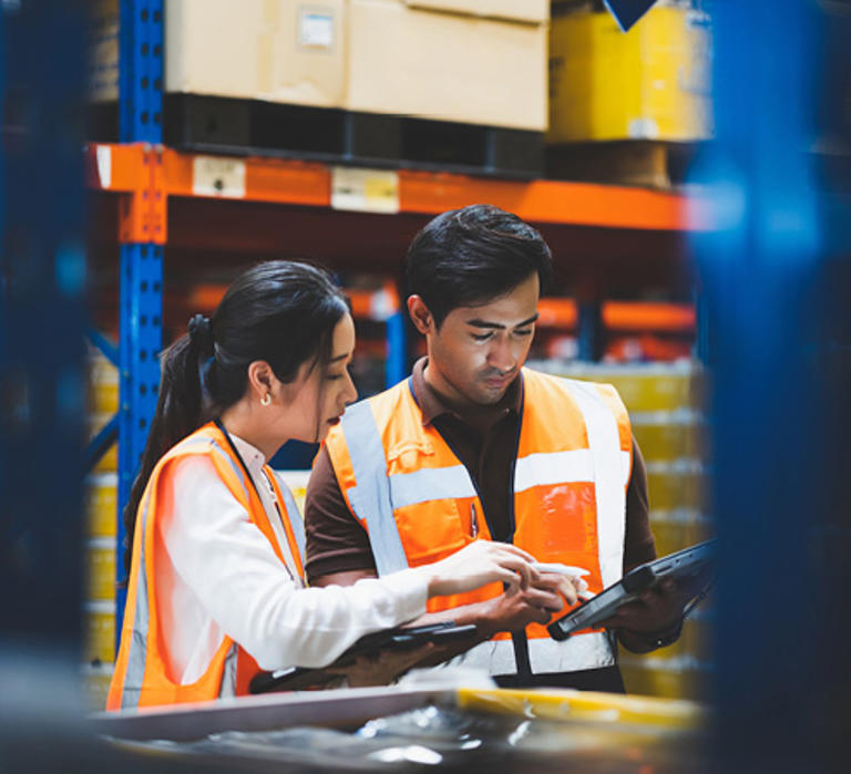 Man and woman in orange vests looking at a tablet inside a warehouse