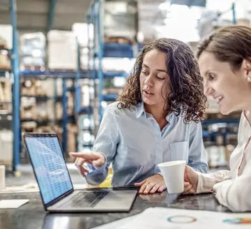 Two women are looking at a laptop together inside of a warehouse