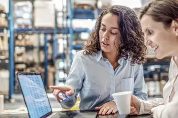 Two women are looking at a laptop together inside of a warehouse